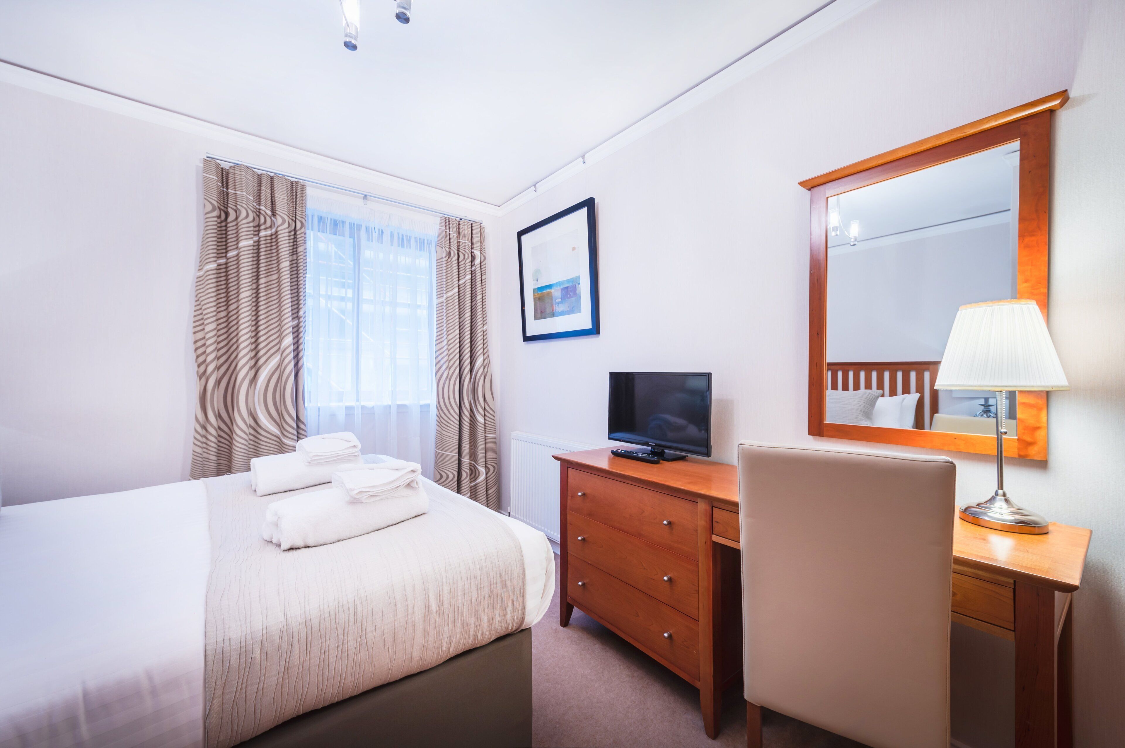A hotel room with a bed, a desk, a chair, a television, a lamp, a mirror, and a window with curtains.