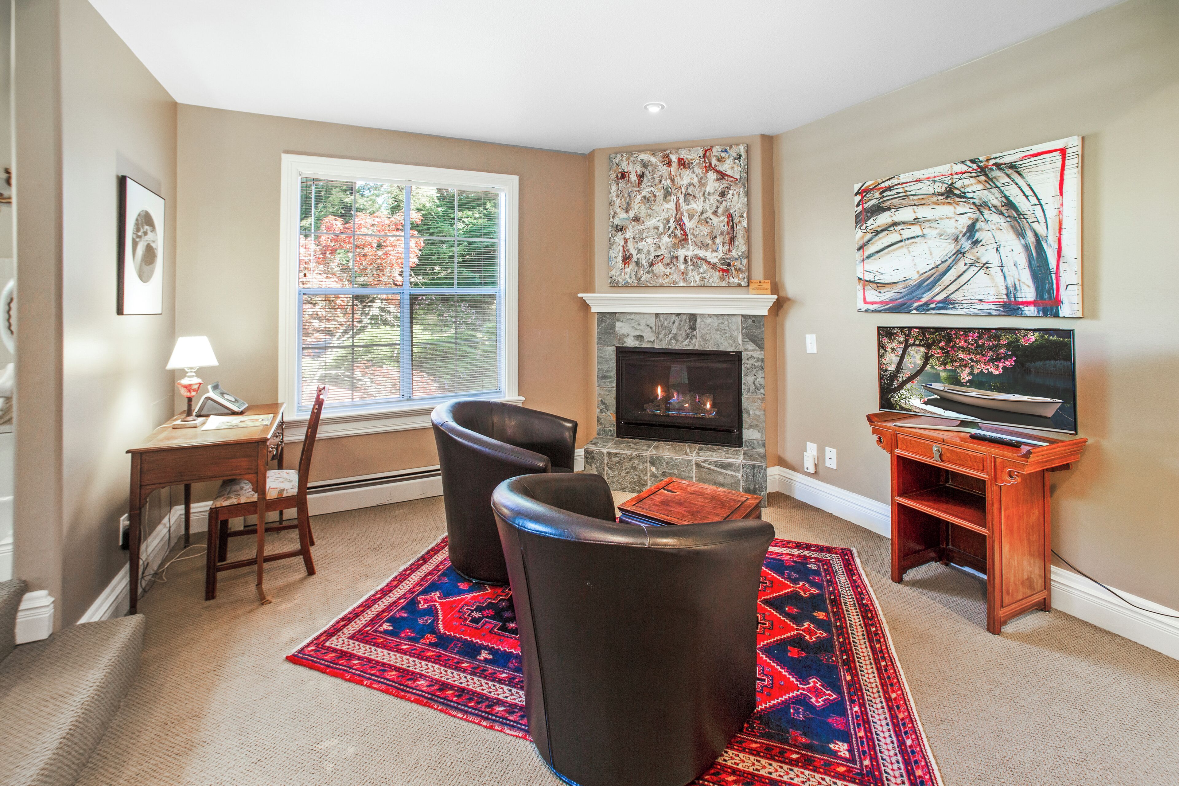 A living room with a fireplace, a desk, two chairs, a rug, and artwork on the walls.