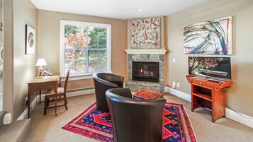 A living room with a fireplace, a desk, two chairs, a rug, and artwork on the walls.
