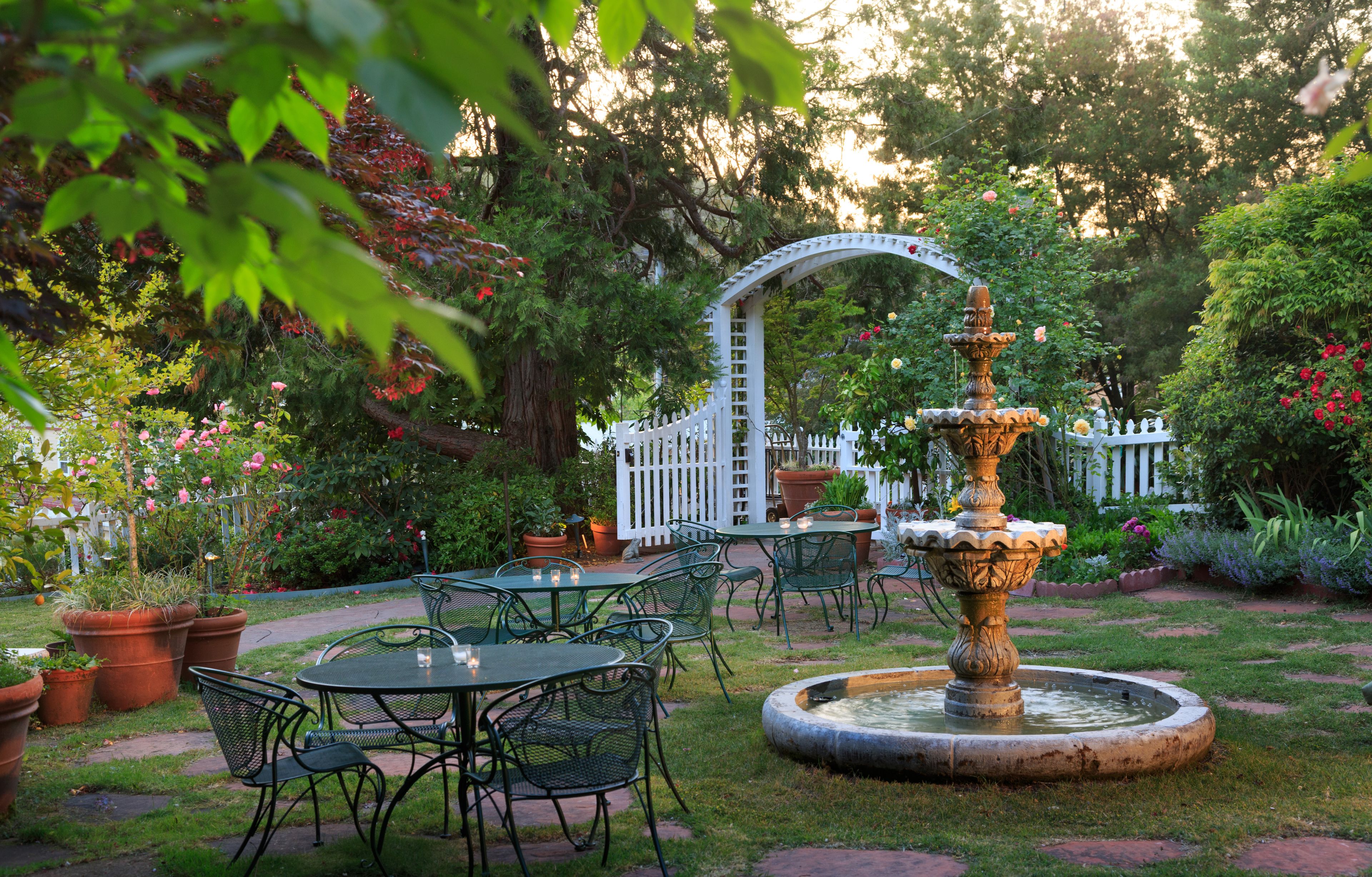 Un jardin agrémenté d’une fontaine, de tables, de chaises et d’une clôture blanche en bois.