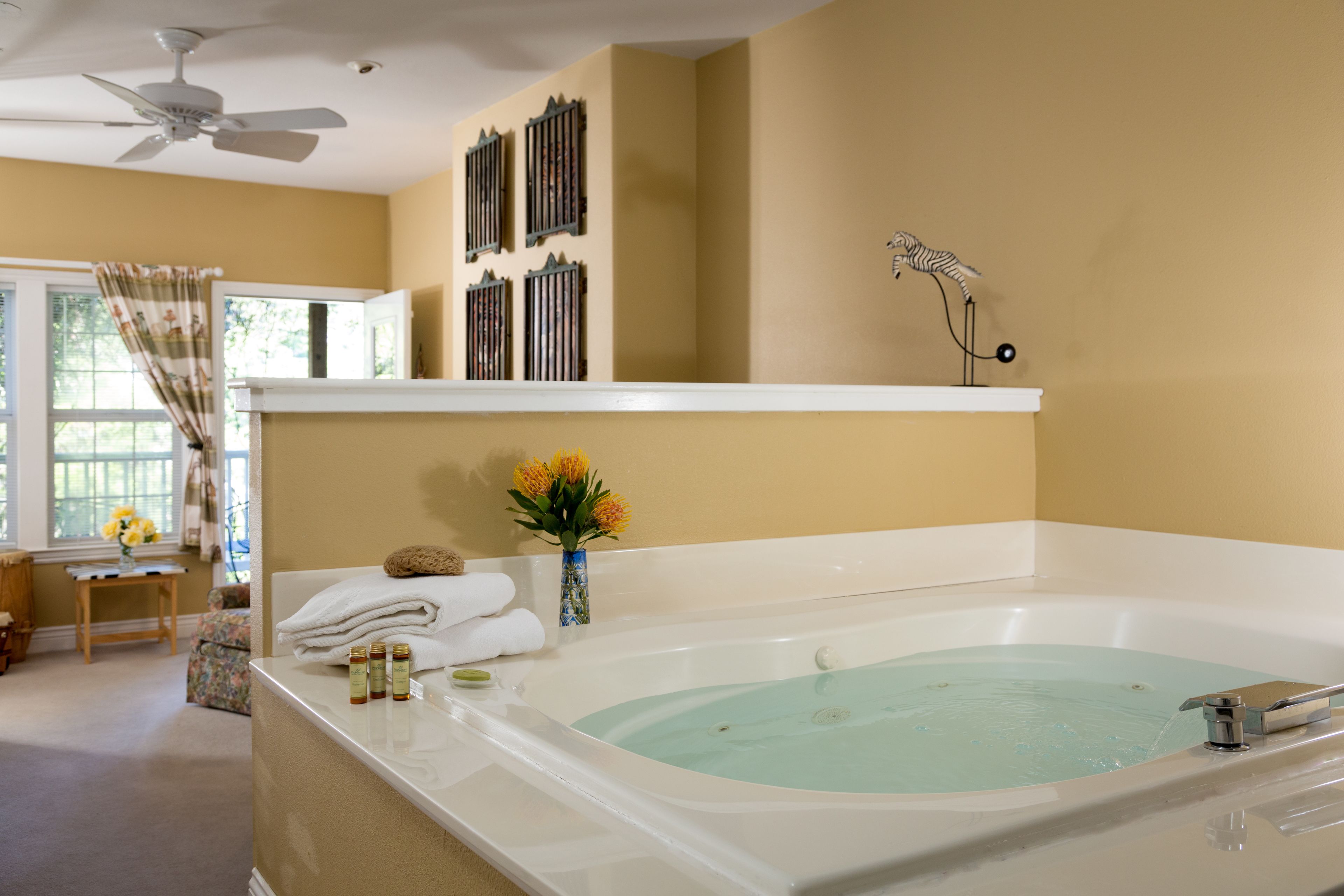 A bathroom with a large bathtub, a ceiling fan, and a window with curtains.