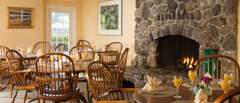 A dining area with wooden chairs and tables, a stone fireplace, and artwork on the walls.