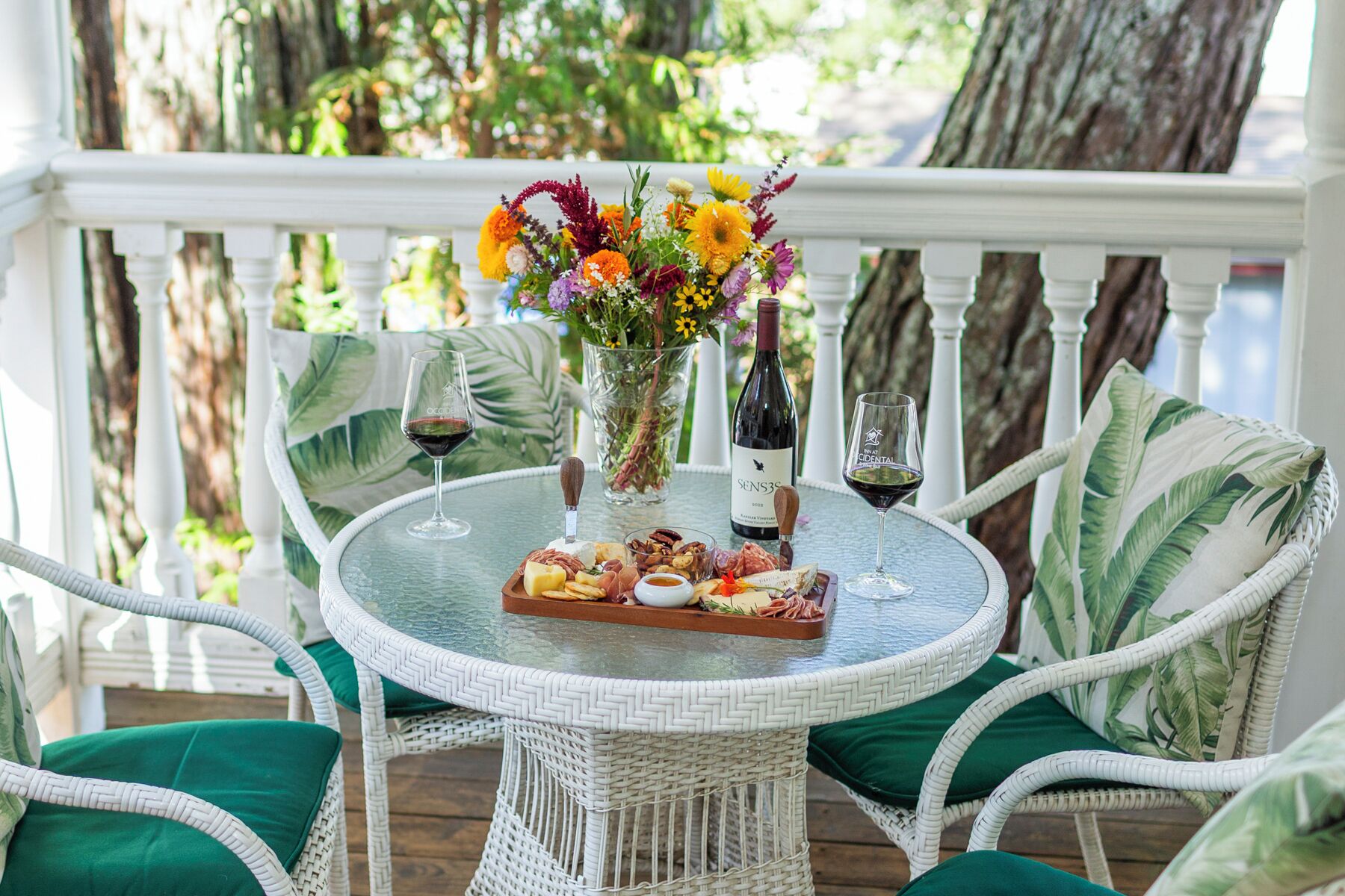 A round glass table with a wine bottle, glasses, a cheese board, and a floral centerpiece on a porch with wicker chairs and a white railing.