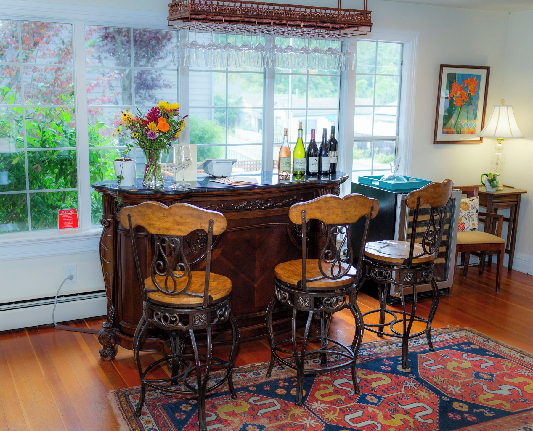 A bar area with a wooden counter, two bar stools, a vase with flowers, and bottles of wine.