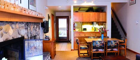 A living room with a stone fireplace, wooden dining table, and chairs. There is a kitchen area with wooden cabinets and a staircase leading to another level.