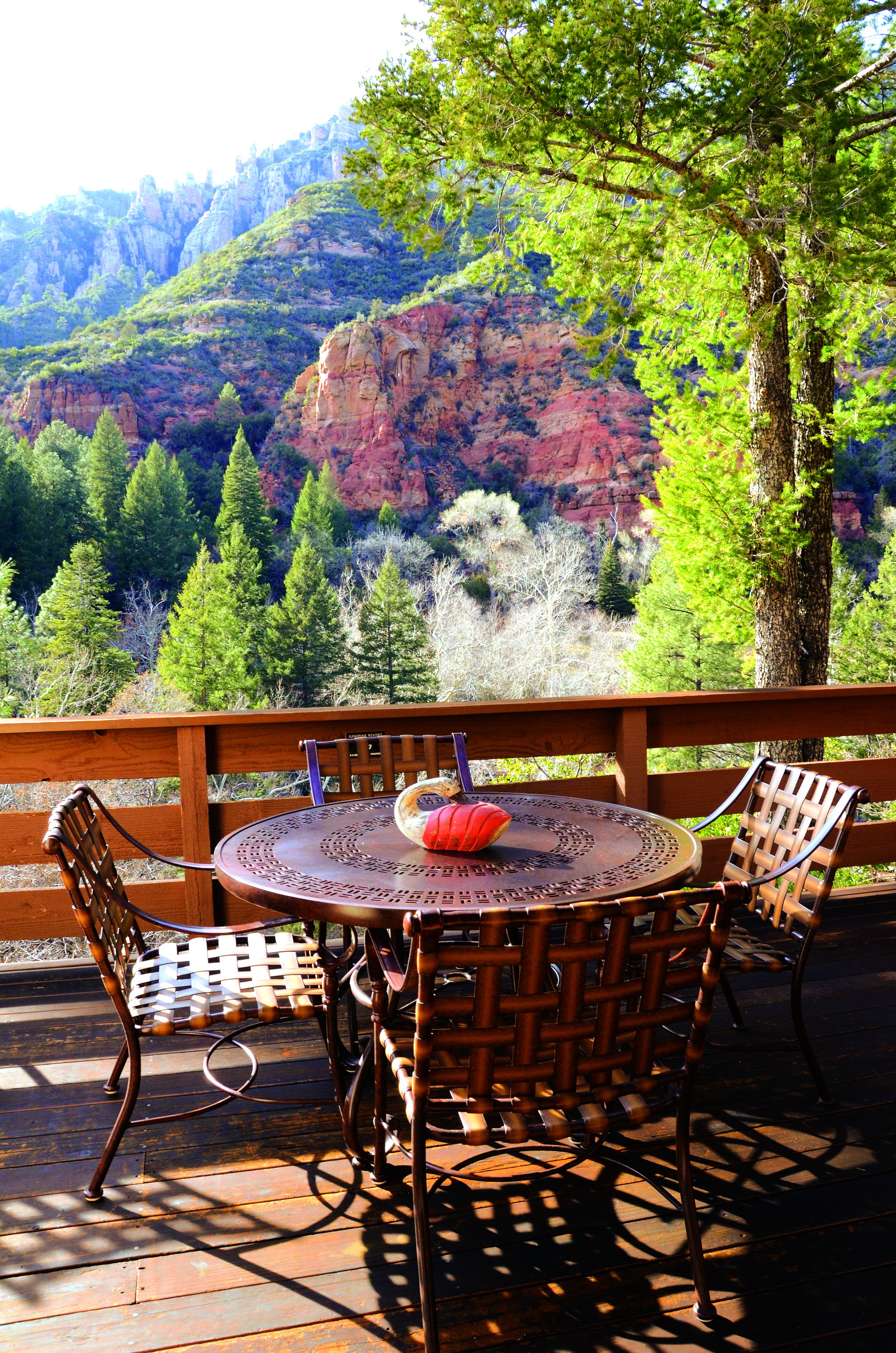 A wooden deck with wicker chairs and a round table, overlooking a mountainous landscape with trees and red rock formations.