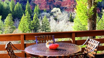A wooden deck with wicker chairs and a round table, overlooking a mountainous landscape with trees and red rock formations.