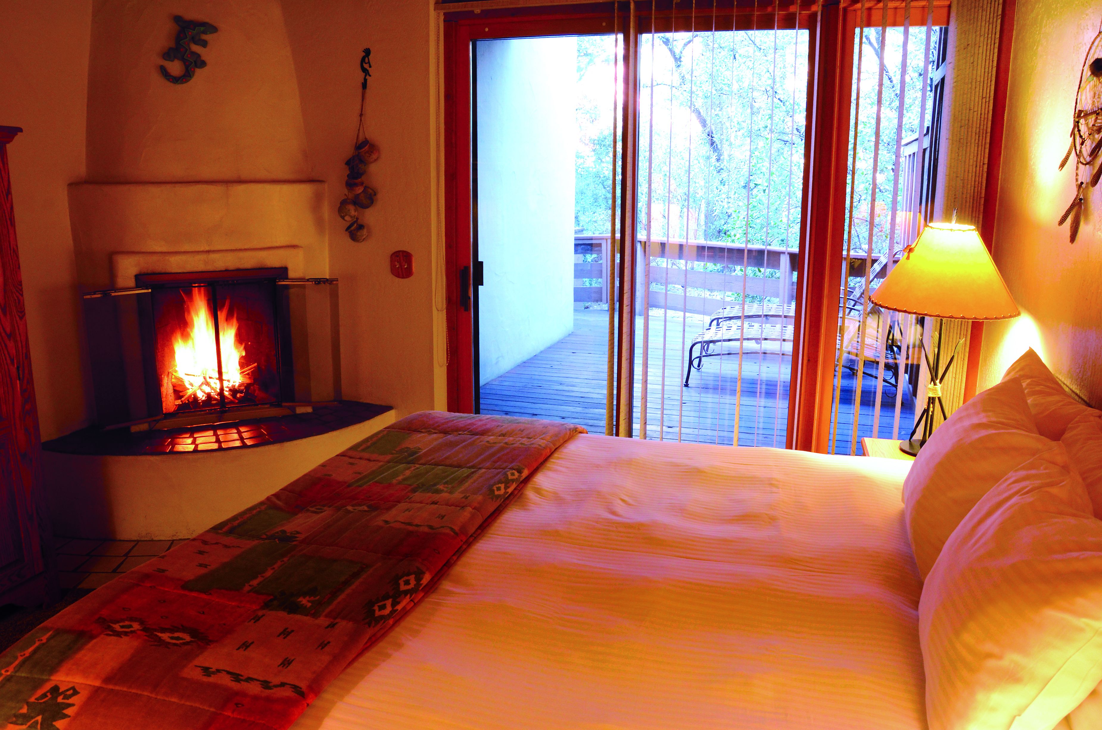 Ein Schlafzimmer mit Kamin, einem Bett mit farbenfroher Decke und Blick auf eine Terrasse und Bäume.