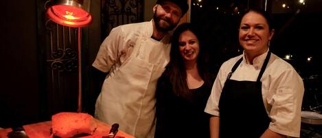 A chef in a white uniform and apron standing next to a woman in a black dress, both smiling in a dimly lit kitchen with a red light and a large cutting board with a knife and a block of meat.