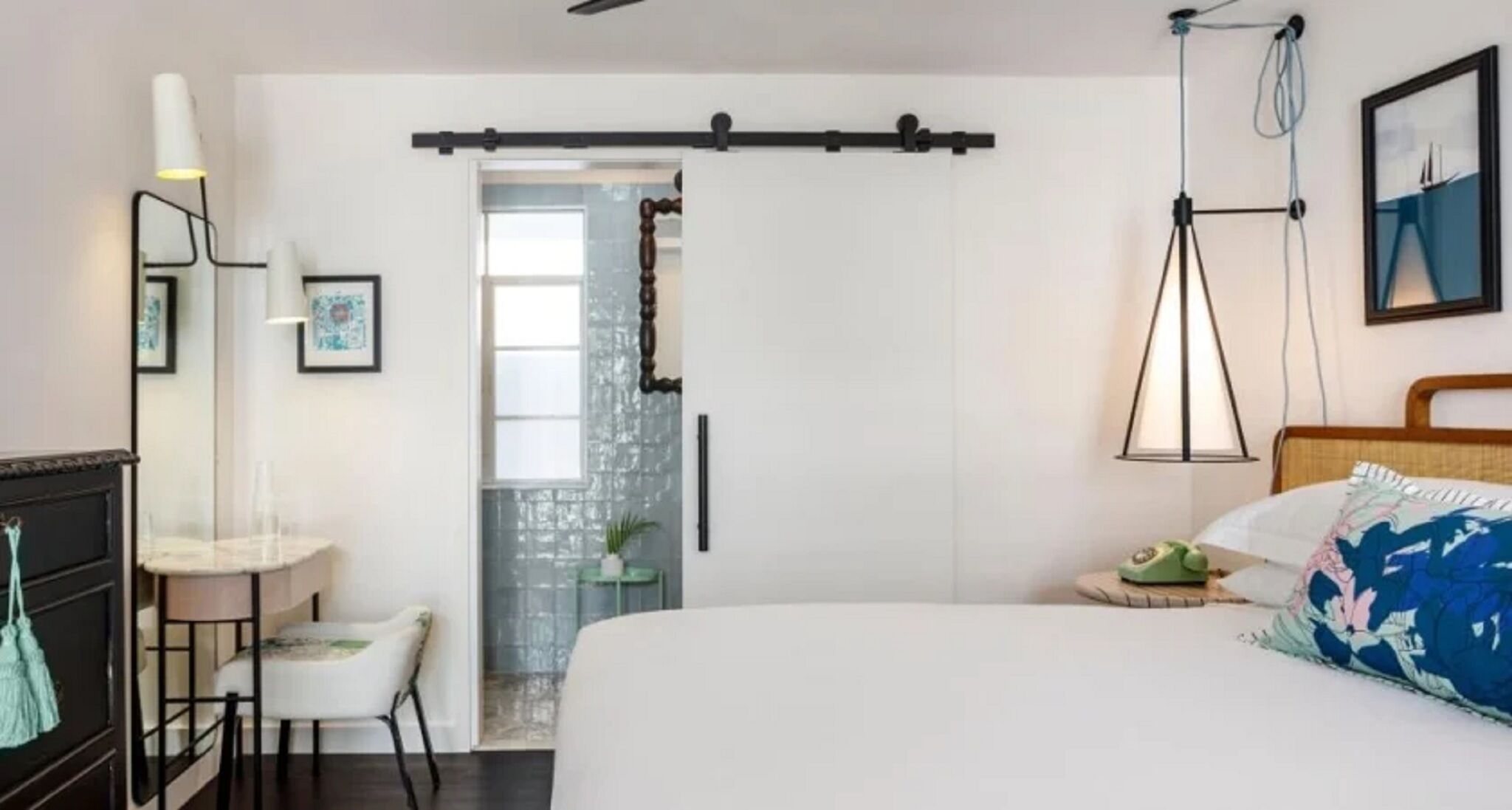 A bedroom with a bed, a desk, a chair, a dresser, a door, and a wall-mounted lamp.