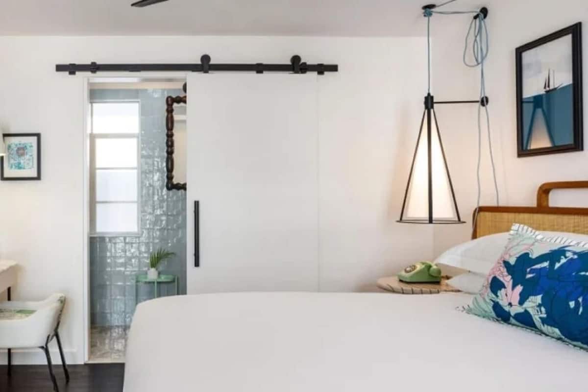A bedroom with a bed, a desk, a chair, a dresser, a door, and a wall-mounted lamp.