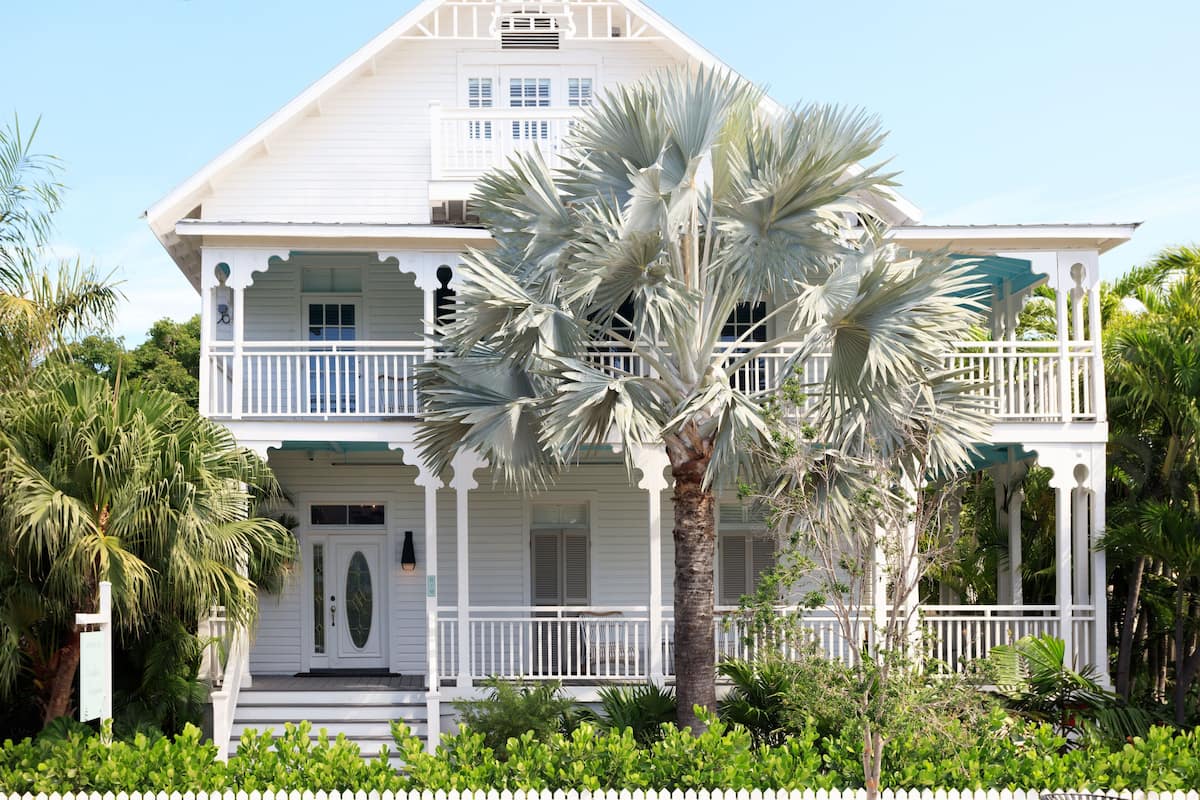A two-story white house with a balcony, surrounded by lush greenery and a white picket fence.