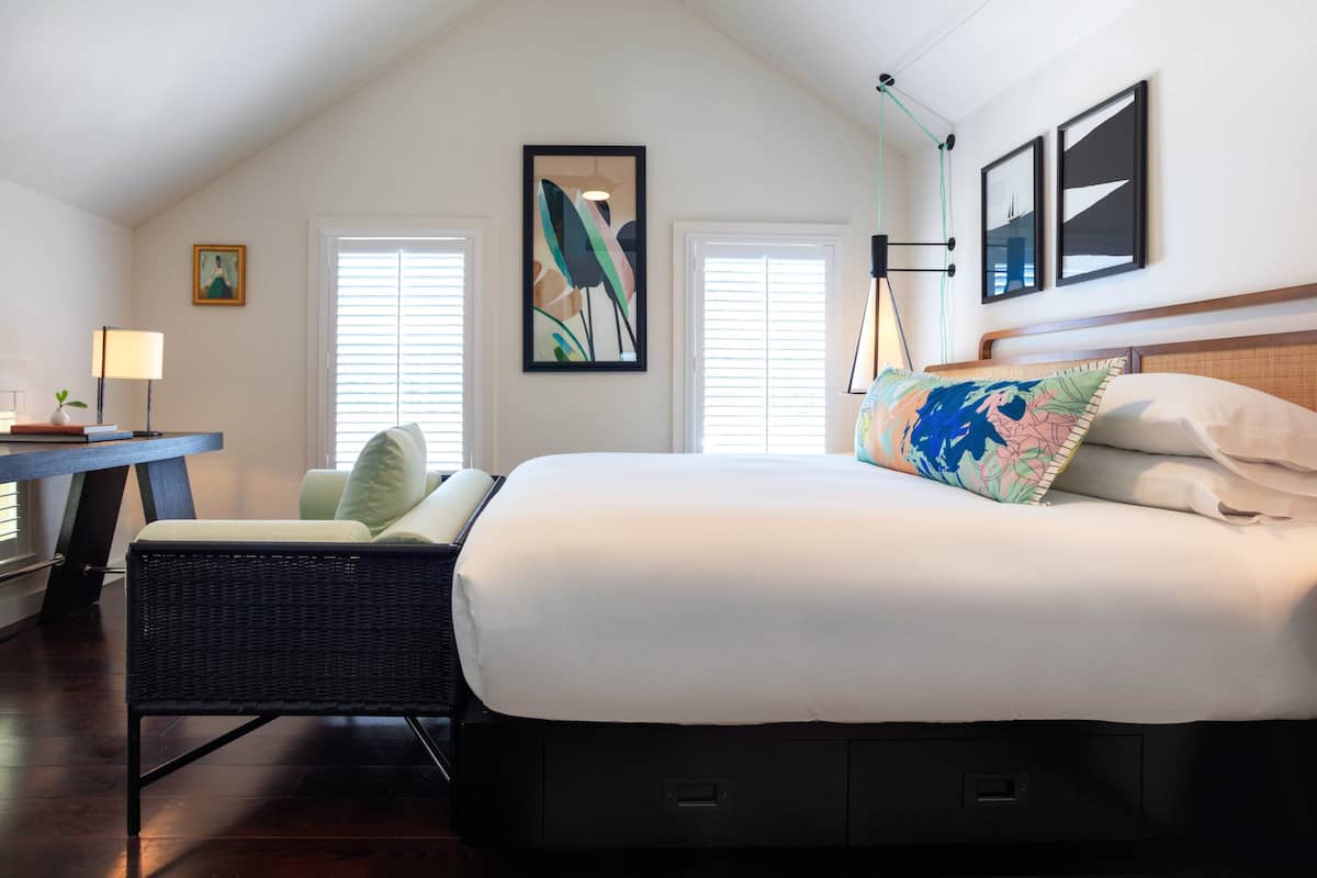 A bedroom with a large bed, a wicker chair, a desk with a lamp, and a painting on the wall.