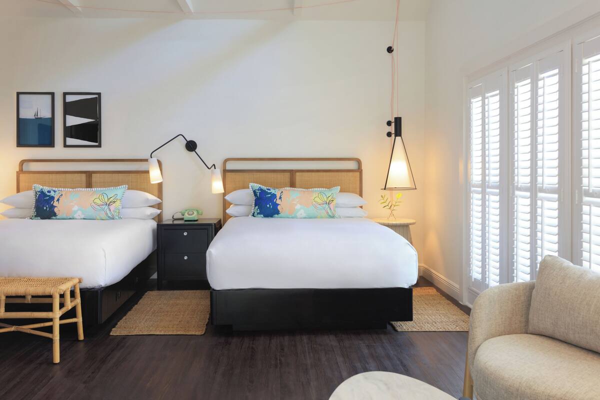 A hotel room with two beds, a wooden headboard, a bedside table with a lamp, a wicker chair, and a large window with shutters.
