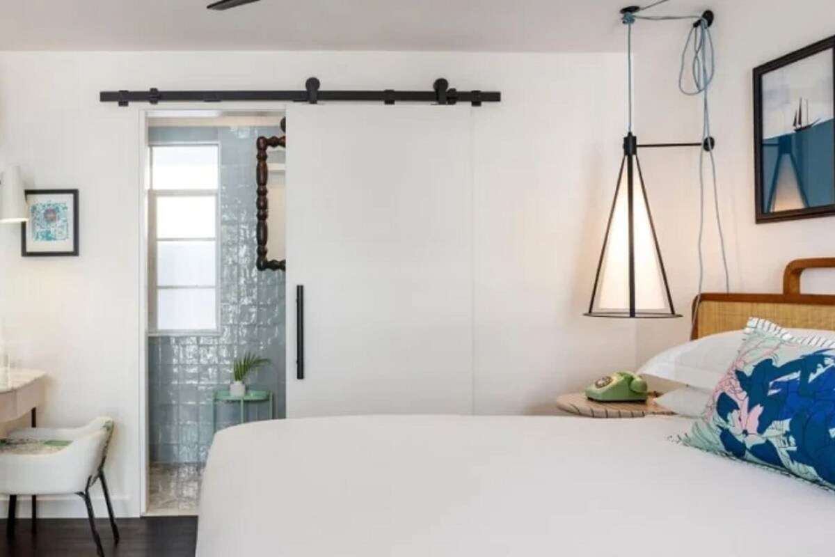 A bedroom with a bed, a desk, a chair, a dresser, a door, and a wall-mounted lamp.