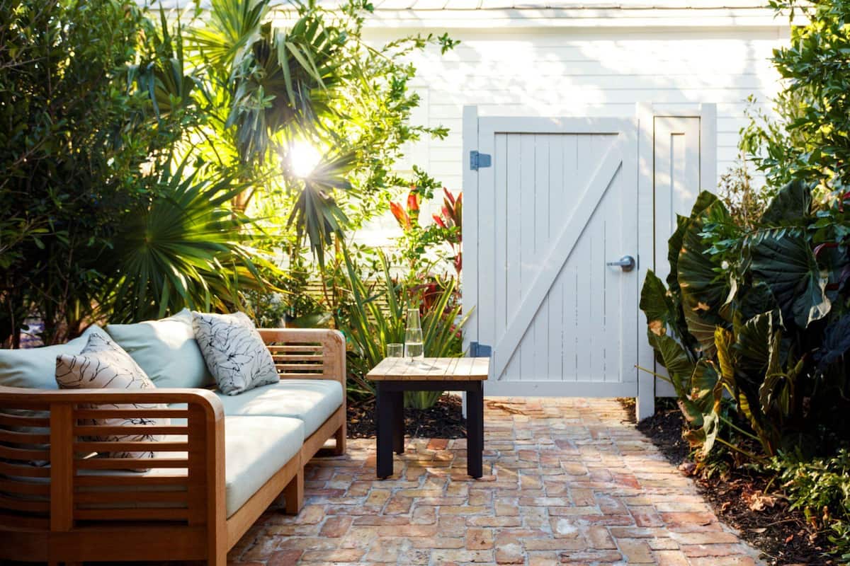 A wooden bench with cushions, a small table with glasses, and a white gate in a garden with various plants.
