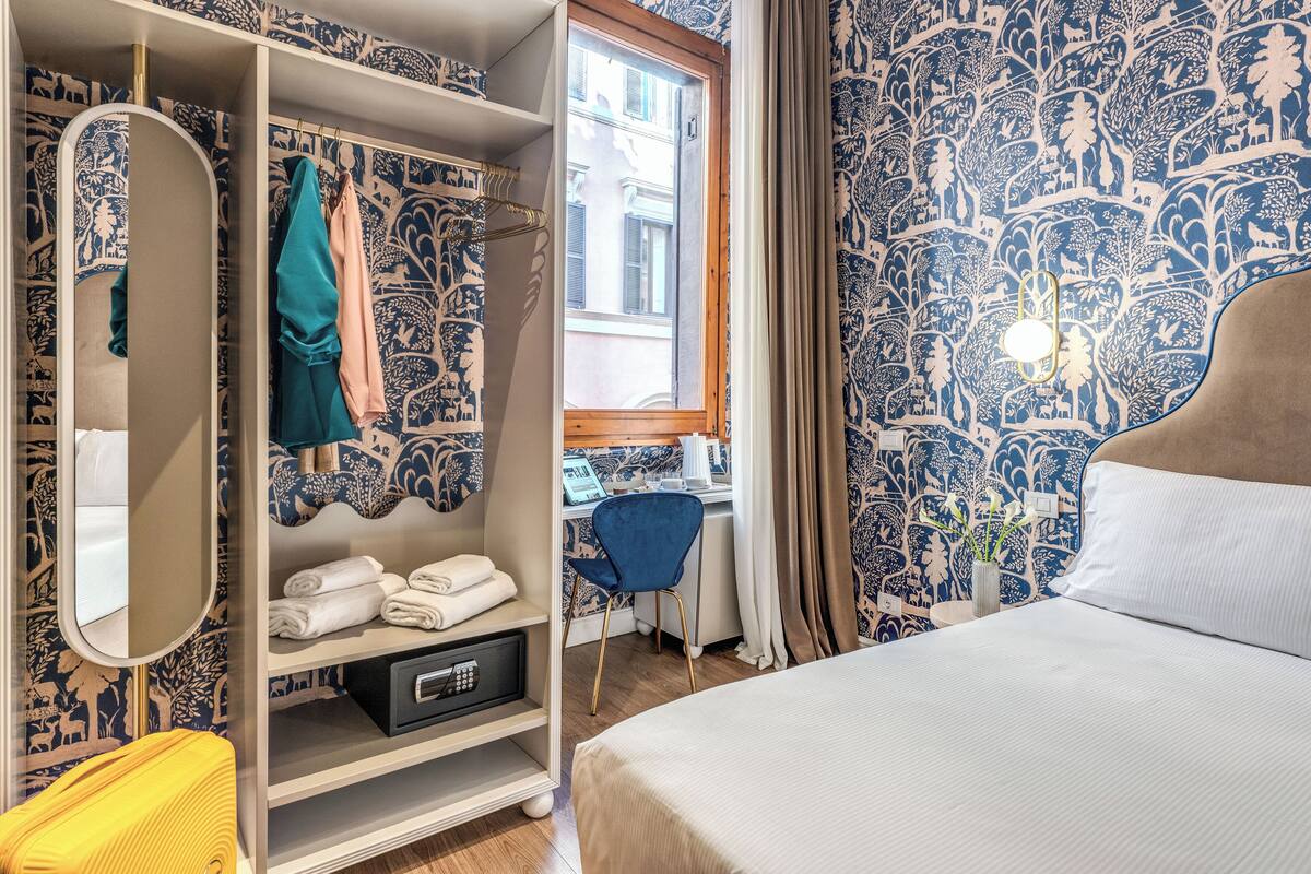Ett hotellrum med en säng, en garderob, ett skrivbord med en stol och ett fönster med utsikt över byggnader.