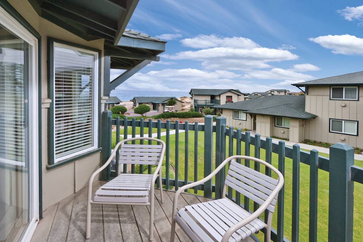 Un balcon avec deux chaises en métal et une terrasse en bois, donnant sur un quartier résidentiel avec des maisons et un ciel dégagé.
