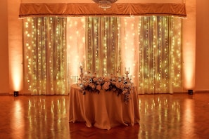 Experience a sophisticated atmosphere in this elegantly set-up room, featuring a draped curtain with string lights, a table with a floral arrangement and candles.