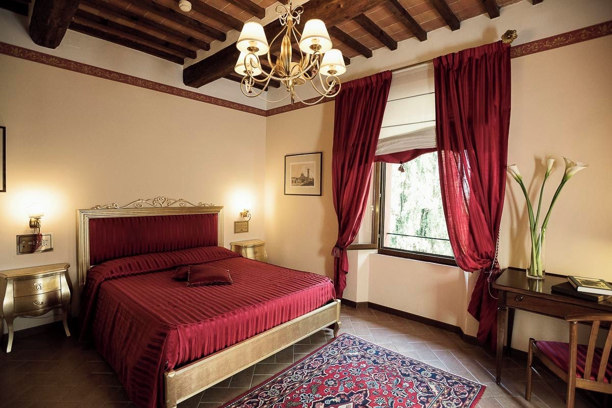 A bedroom with a large bed, a chandelier, a window with red curtains, and a nightstand with a lamp.