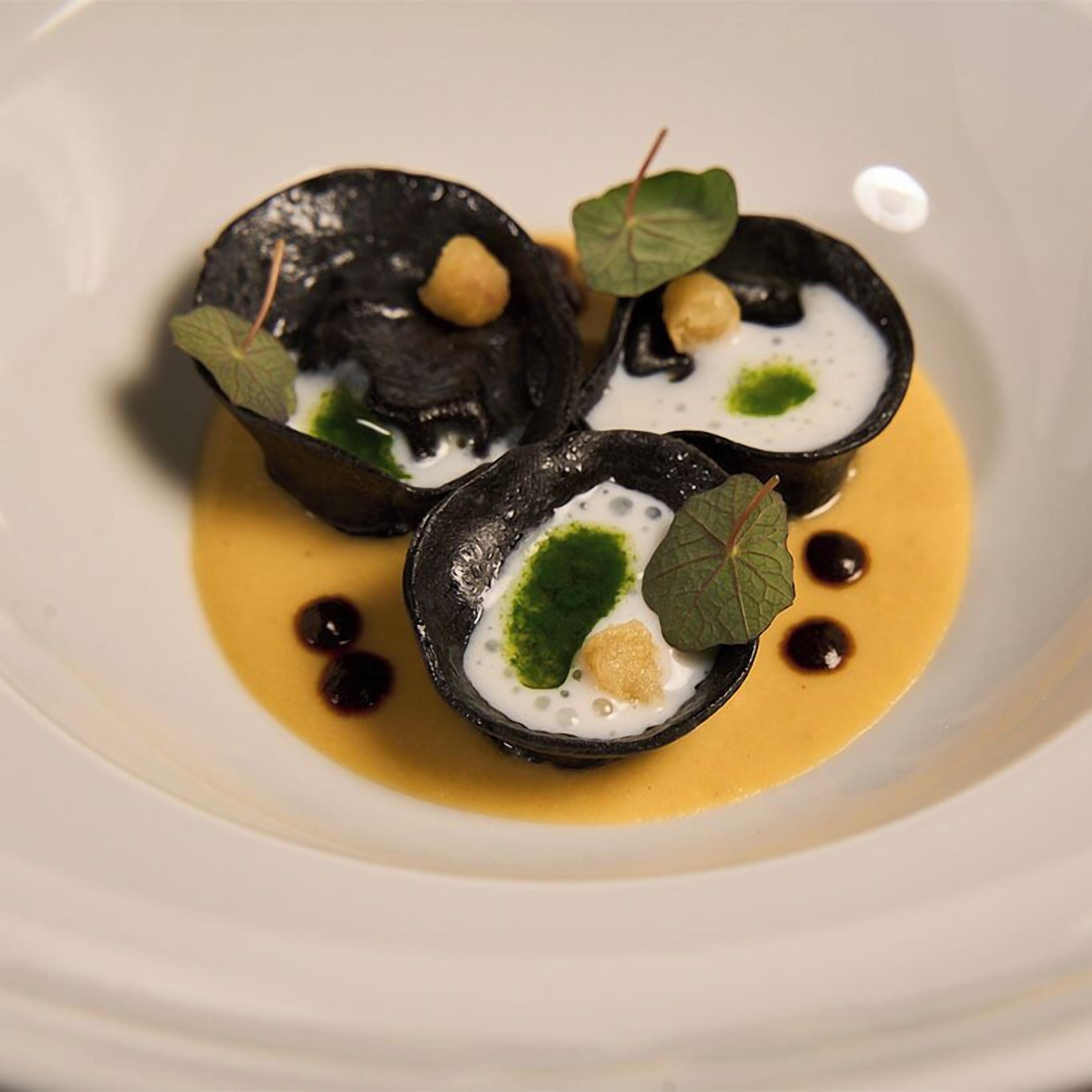 A white plate with three black bowls containing a creamy substance, garnished with green leaves and small brown items.