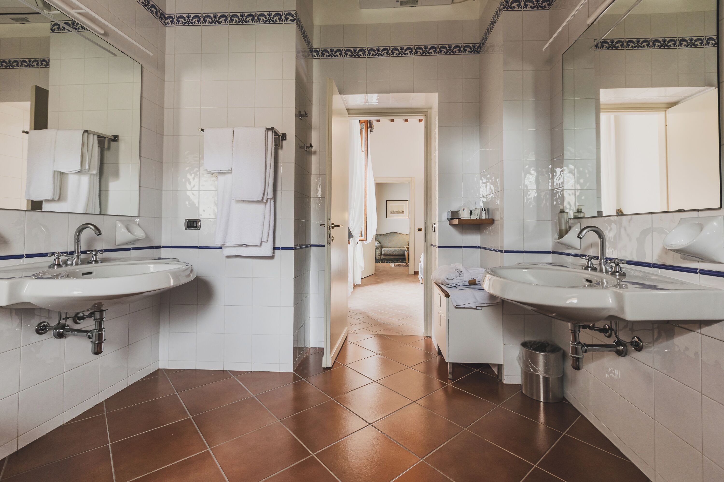 Une salle de bain avec deux lavabos, des murs recouverts de carreaux blancs et un sol carrelé de couleur marron.
