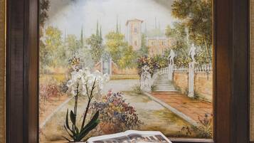 Un cuadro enmarco con una escena de jardín, una planta en maceta y un libro abierto sobre una superficie de madera.