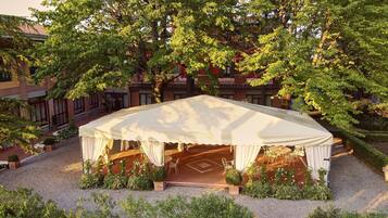 Een overdekt buitenzitgedeelte met tafels en stoelen, omringd door grind en begroeiing, met gebouwen op de achtergrond.