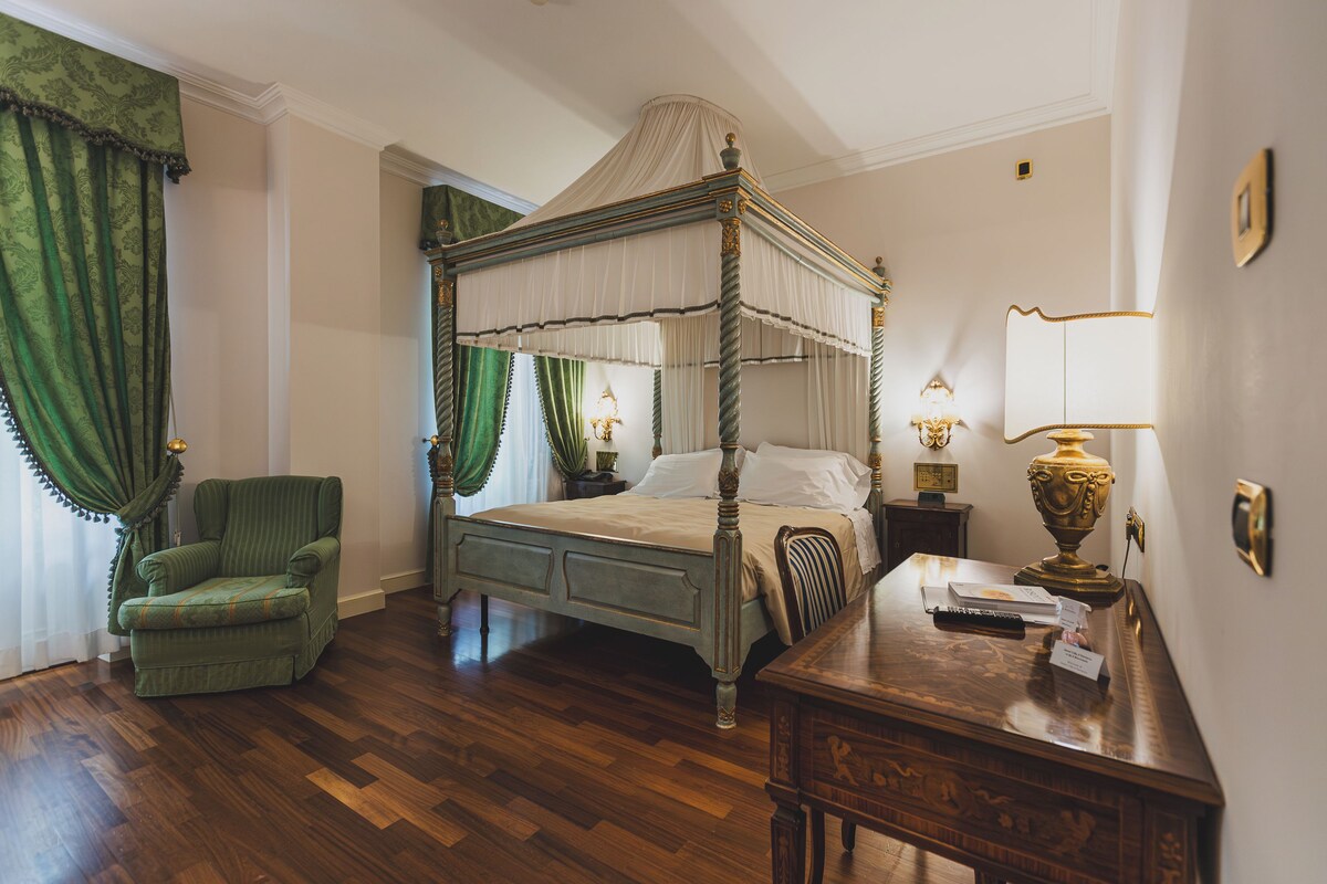 A four-poster bed with a canopy, a green upholstered chair, a wooden desk with a lamp, and a nightstand with a lamp.