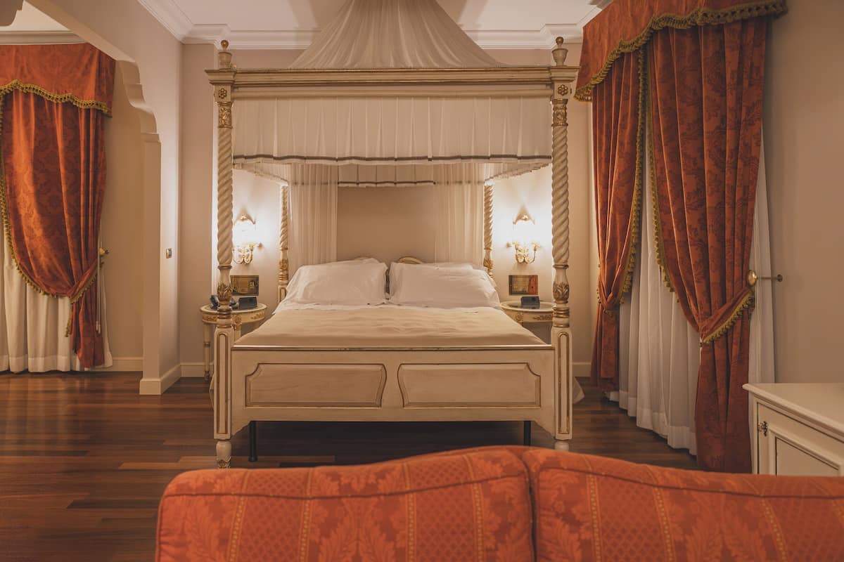 Ein Bett mit Baldachin, an beiden Seiten Nachttische, ein Spiegel über dem Bett und ein kleiner Tisch mit einer Lampe.