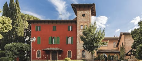 Un edificio rojo con contraventanas verdes y una torre, rodeado de árboles y un camino de grava.