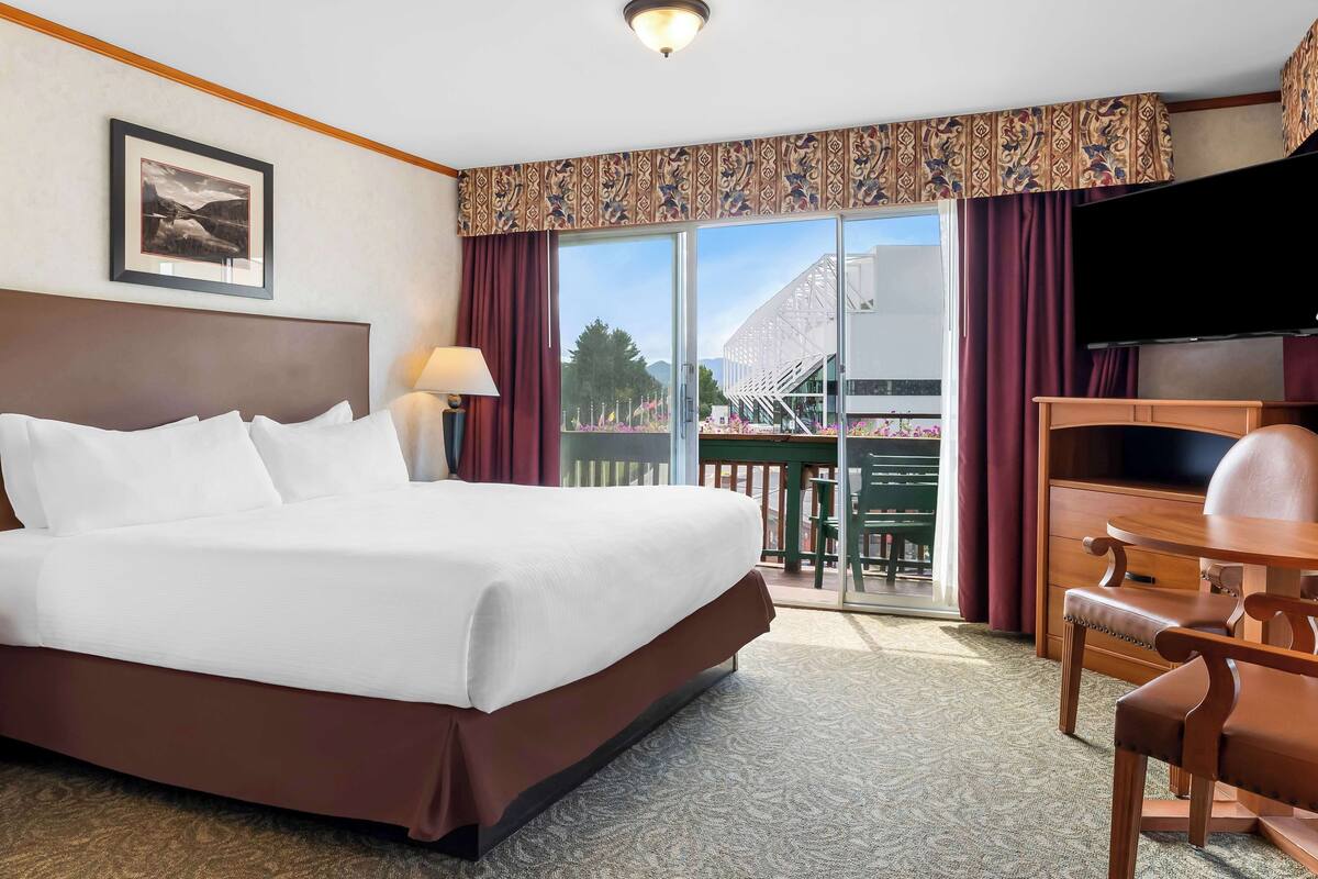 A hotel room with a large bed, a desk with a chair, a television, and a balcony with a view of a building and trees.