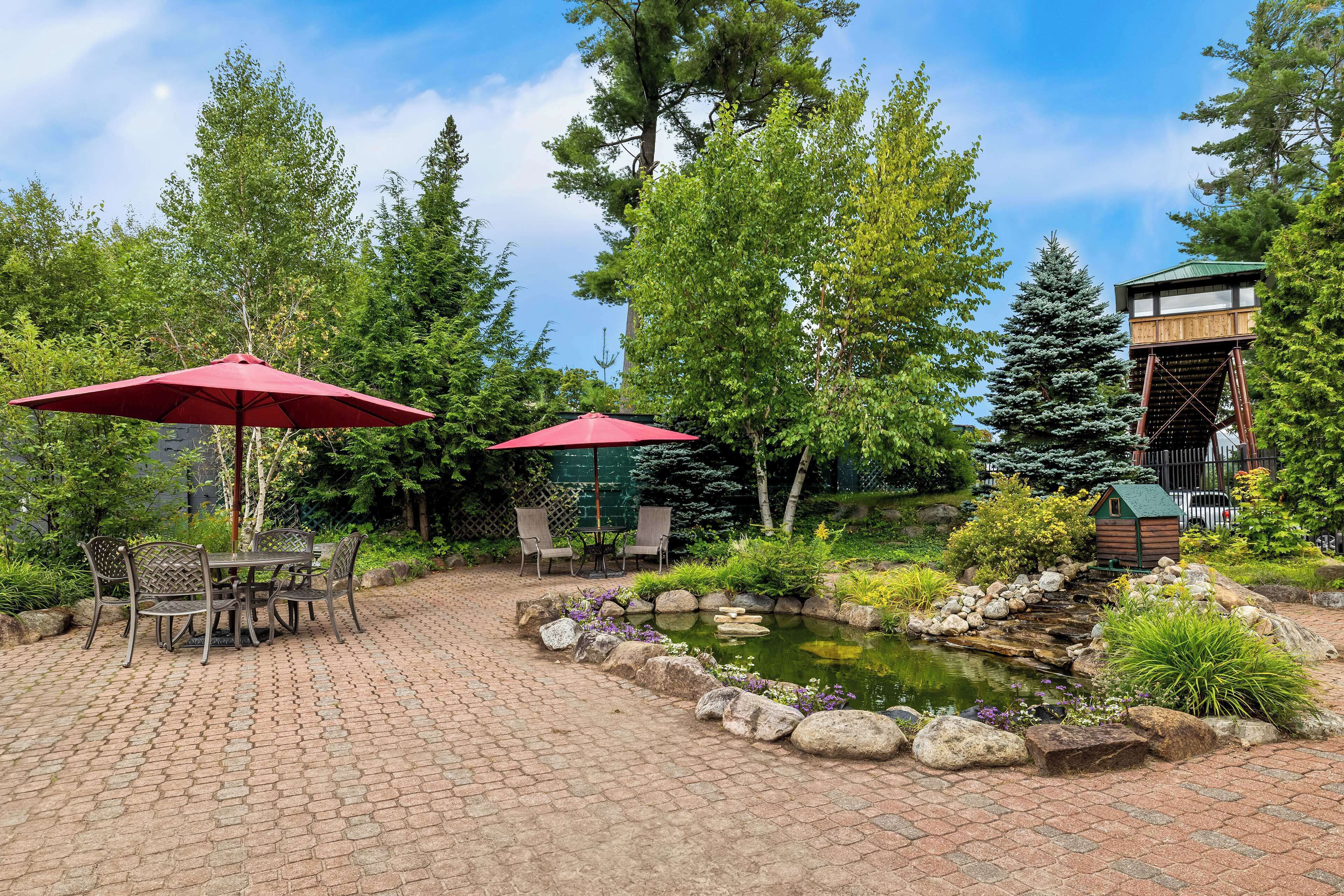 A patio area with a brick pathway, two red umbrellas, a table with chairs, and a small pond with rocks and plants.