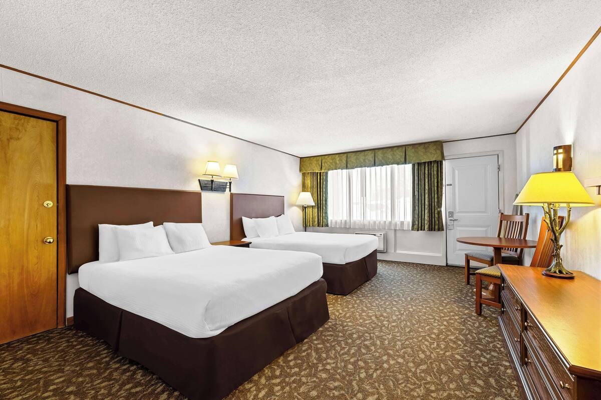 A hotel room with two beds, a desk, a chair, and a lamp.