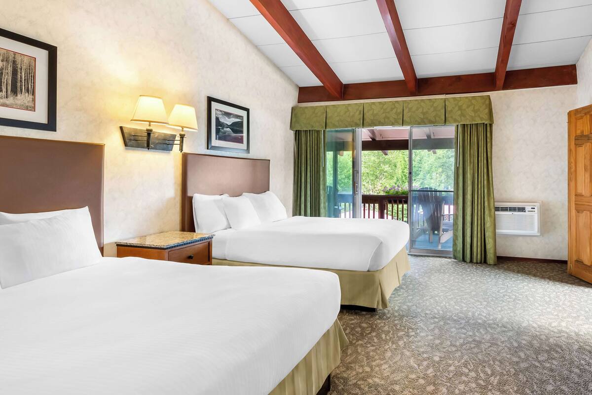 A hotel room with two beds, a nightstand, a lamp, a framed picture, a window with curtains, and a view of greenery outside.