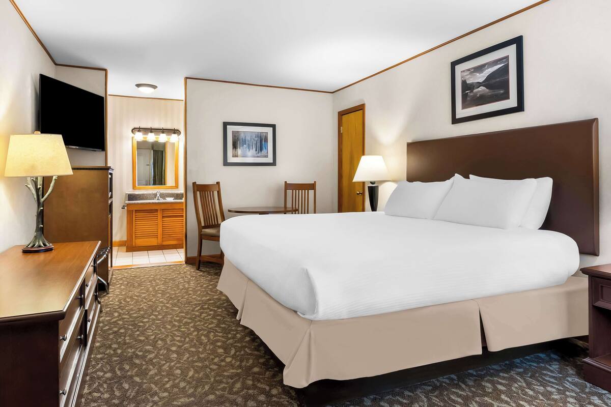 A hotel room with a large bed, a desk, a chair, a vanity with a mirror, and a television.