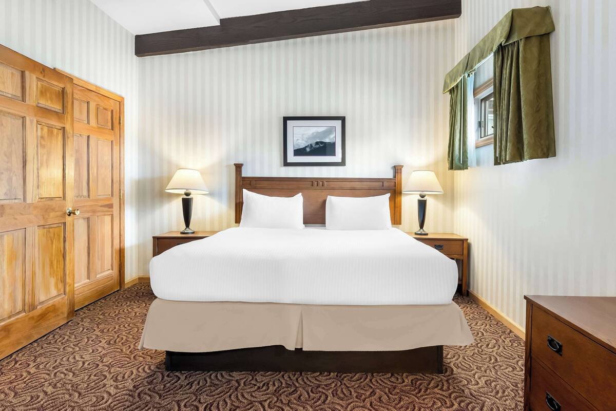 A neatly made bed with a wooden headboard, two bedside tables with lamps, a framed picture on the wall, and a patterned carpet.