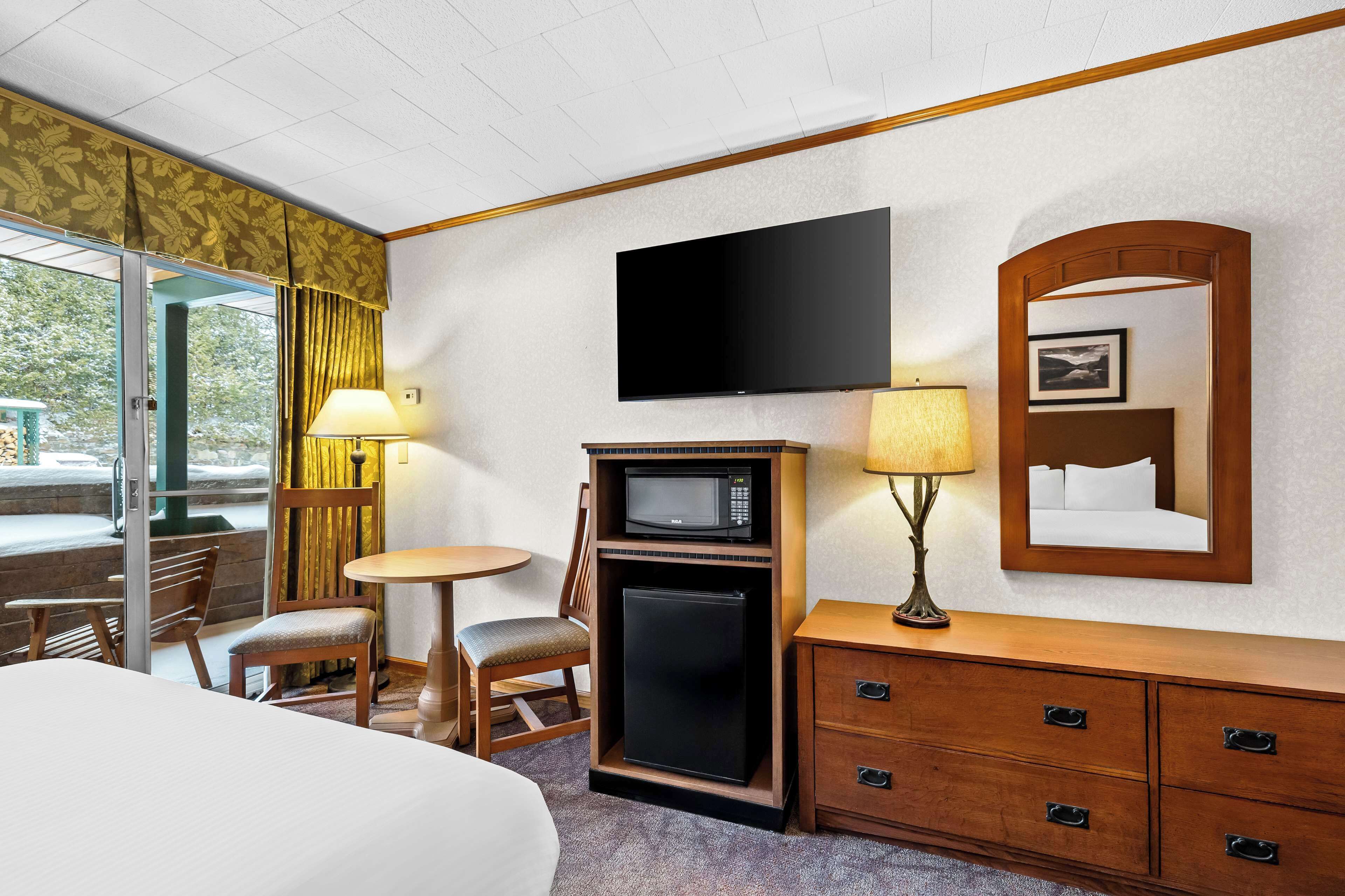 A hotel room with a bed, a desk with a lamp, a microwave, a refrigerator, a dresser, and a television mounted on the wall.