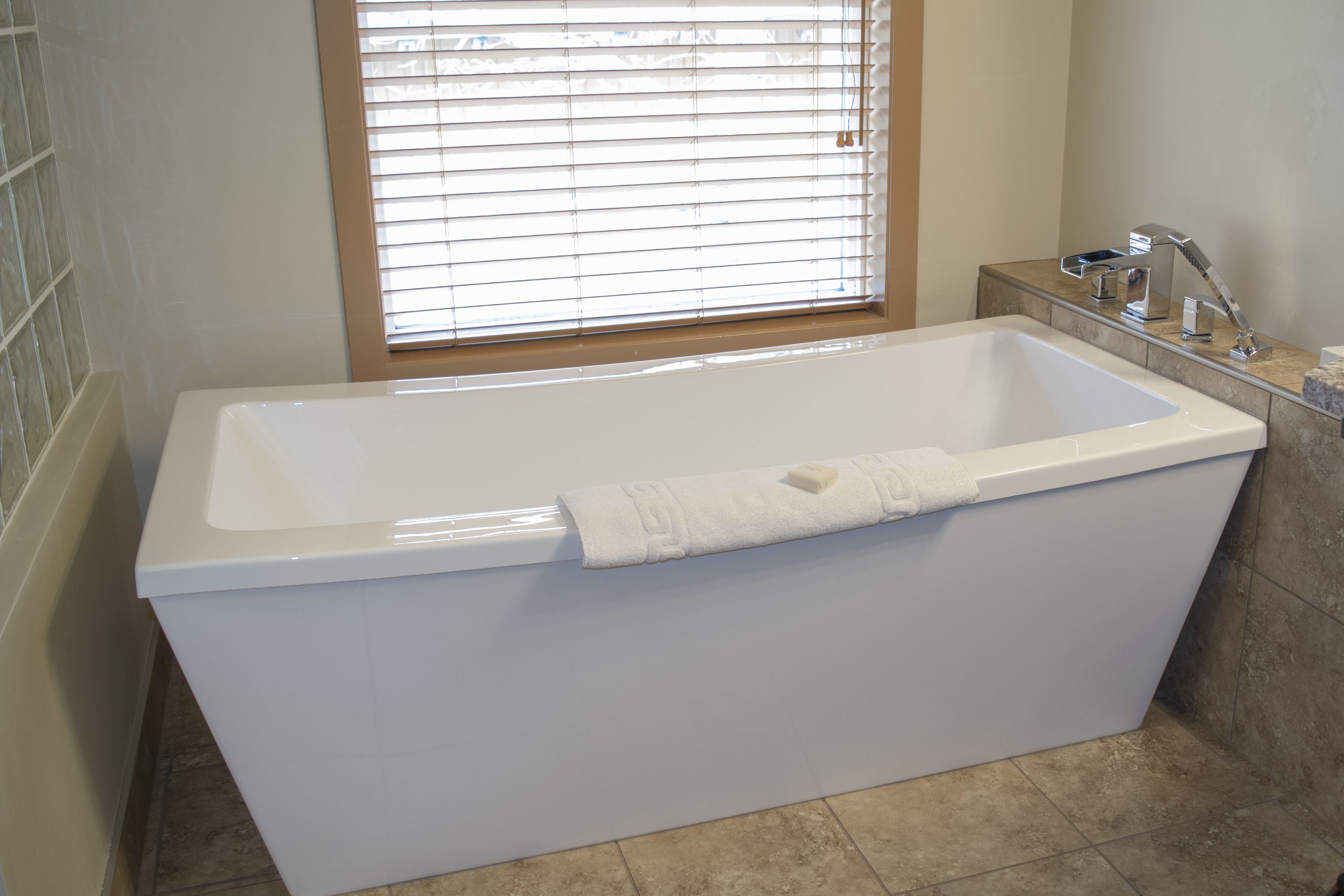 Une salle de bain moderne avec une baignoire rectangulaire, une fenêtre avec des stores et un petit plan de toilette avec un robinet.