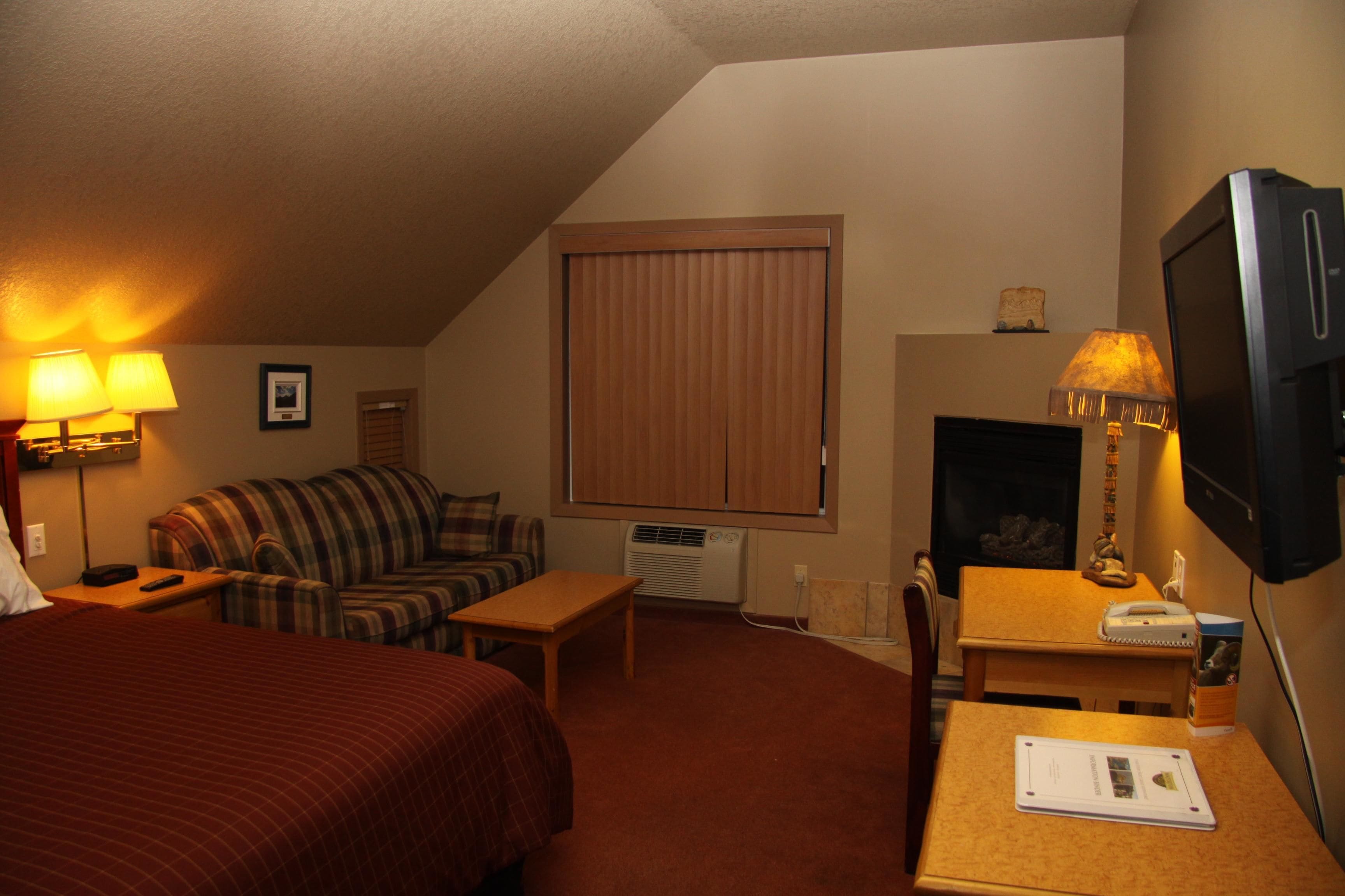 A hotel room with a bed, a sofa, a table, a desk, a TV, and a fireplace.