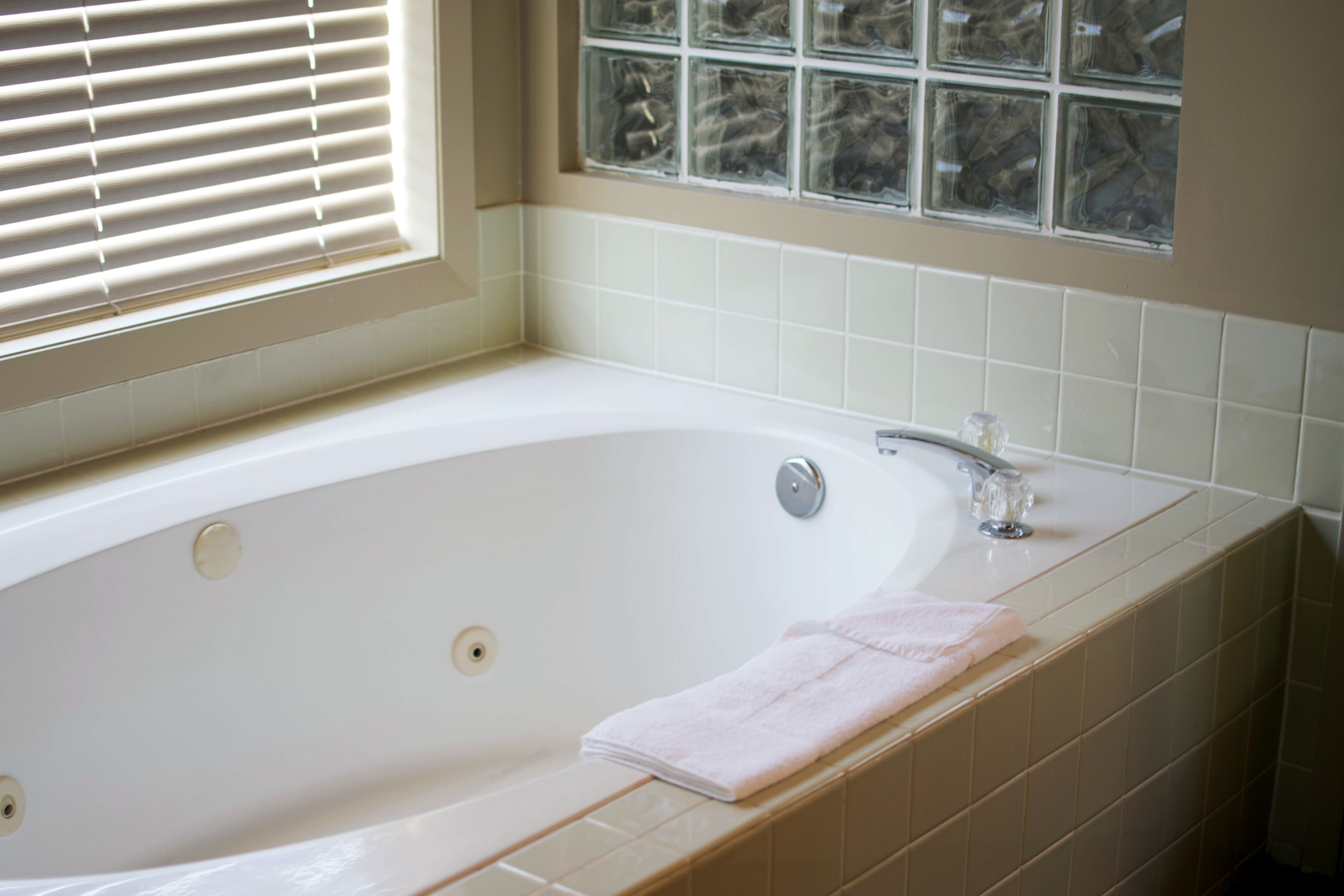 A bathroom with a bathtub, a window with blinds, and a tiled wall.
