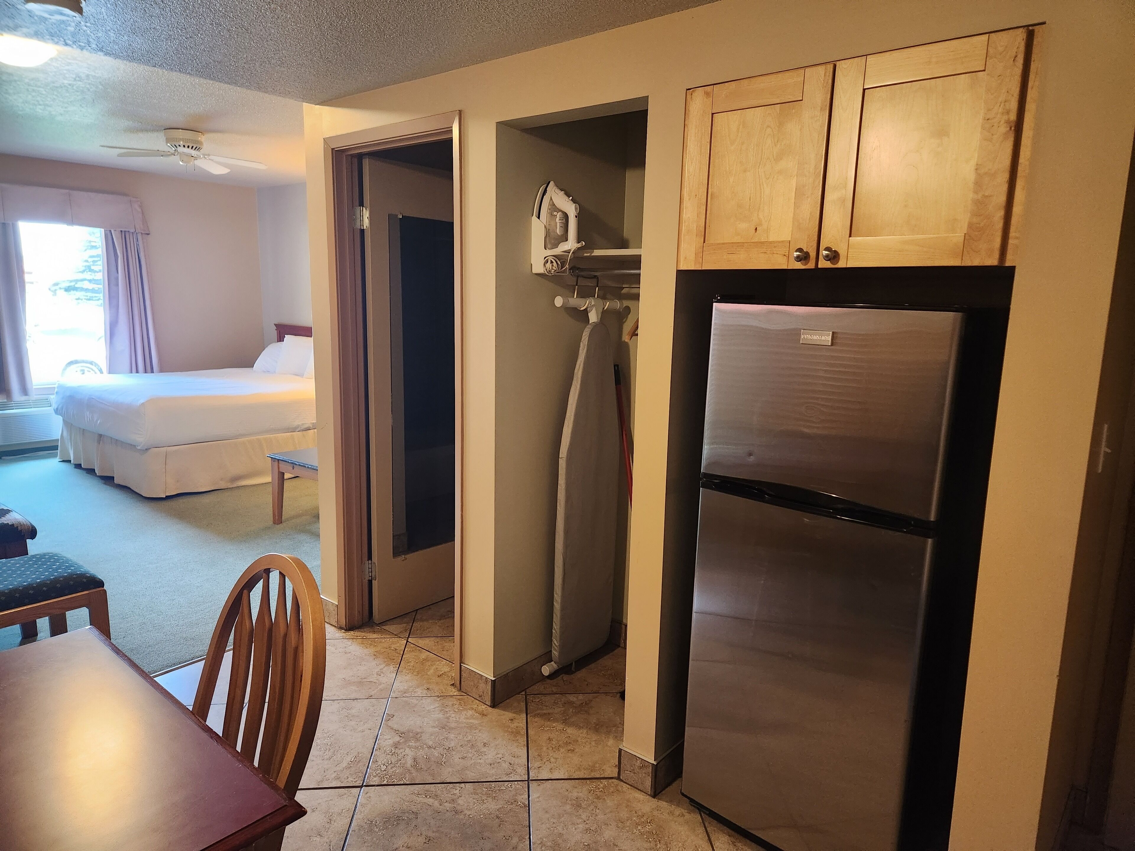 A compact hotel room with a bed, a dining table, and a kitchenette area including a refrigerator and wooden cabinets.