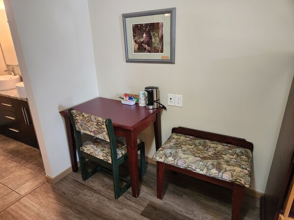 Un pequeño comedor con una mesa y dos sillas, un banco, un cuadro enmarcado en la pared y una cafetera sobre la mesa.