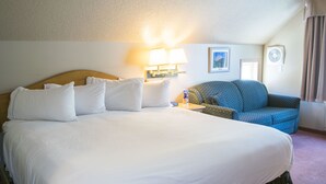 Une chambre d’hôtel avec un grand lit, deux lampes de chevet, une petite table, un fauteuil à motifs bleus et un ventilateur fixé au mur.