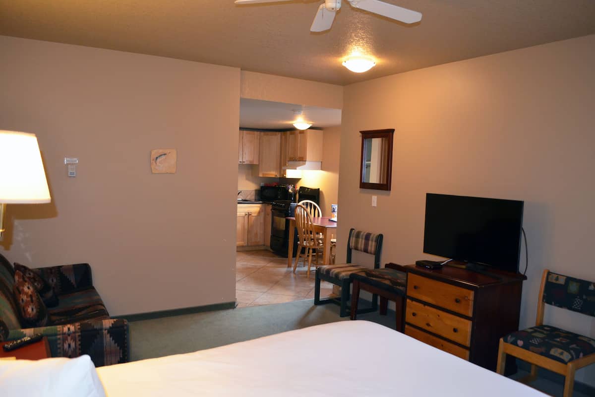 Habitación de hotel con cama, escritorio, silla, cómoda, televisión y una kitchenette al fondo.