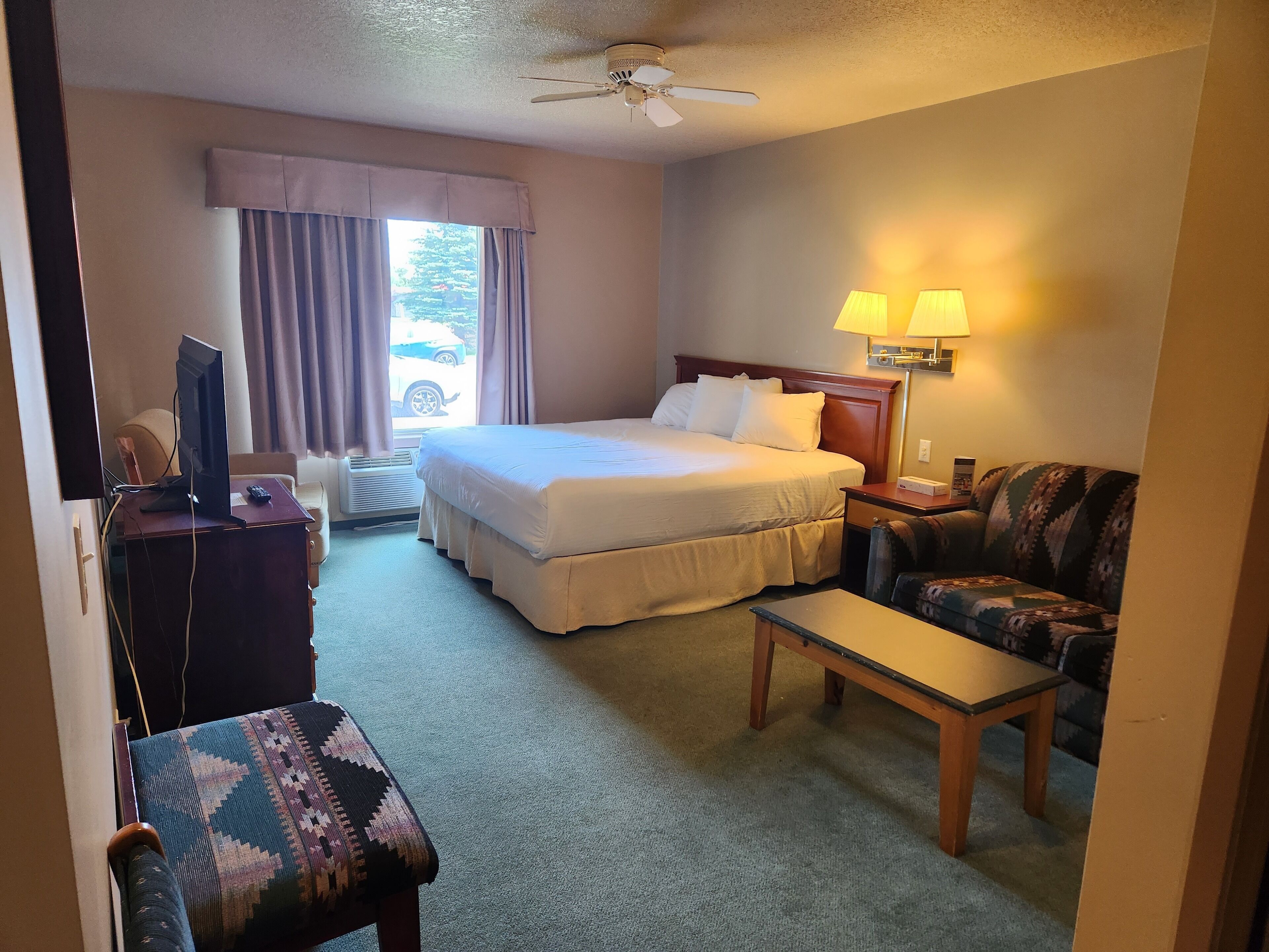 A hotel room with a bed, a desk with a computer, a chair, a sofa, a ceiling fan, and a window with curtains.