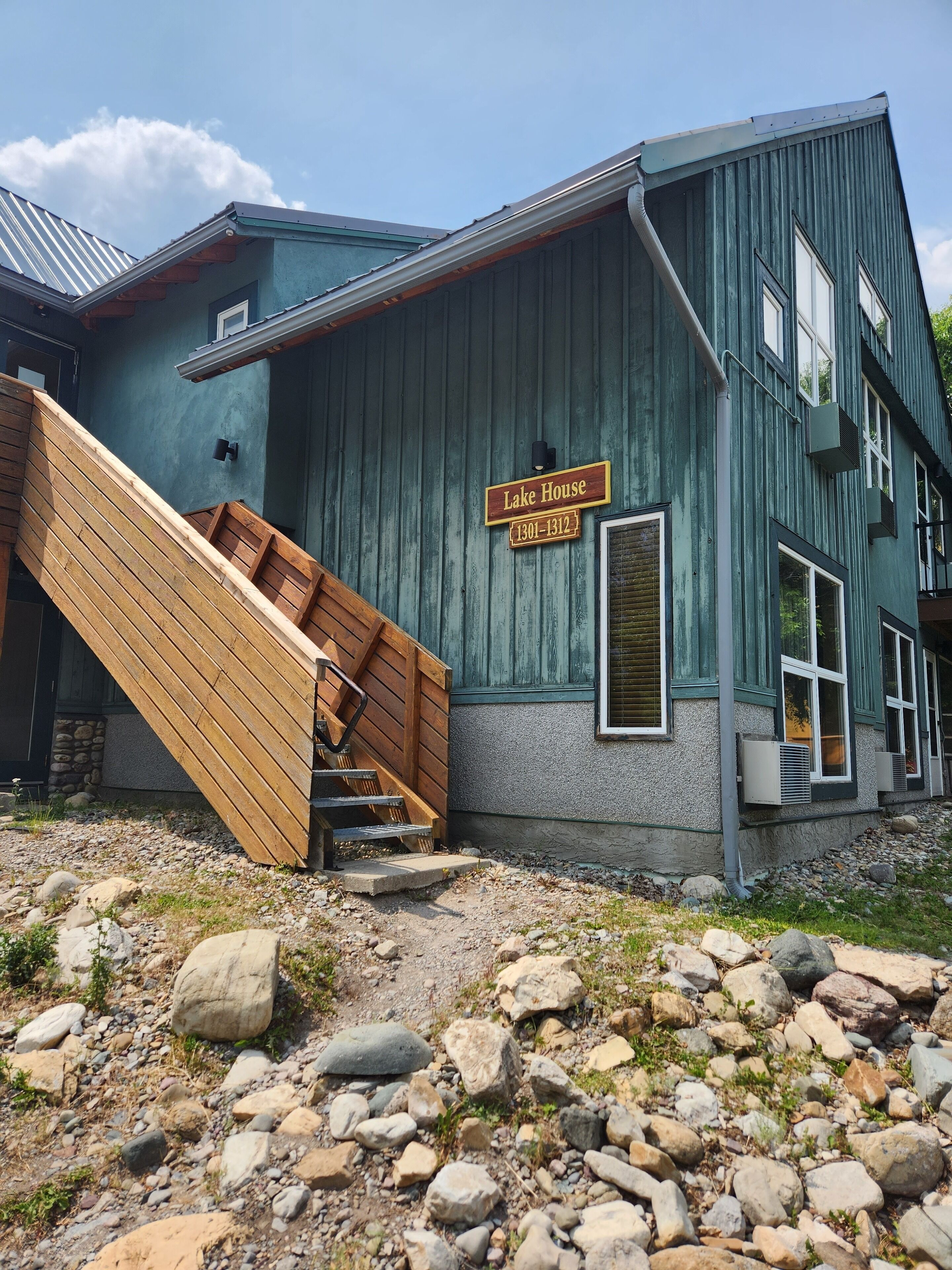 A rustic building with a wooden staircase leading up to a door marked "Lake House" and phone numbers "1301-1312".