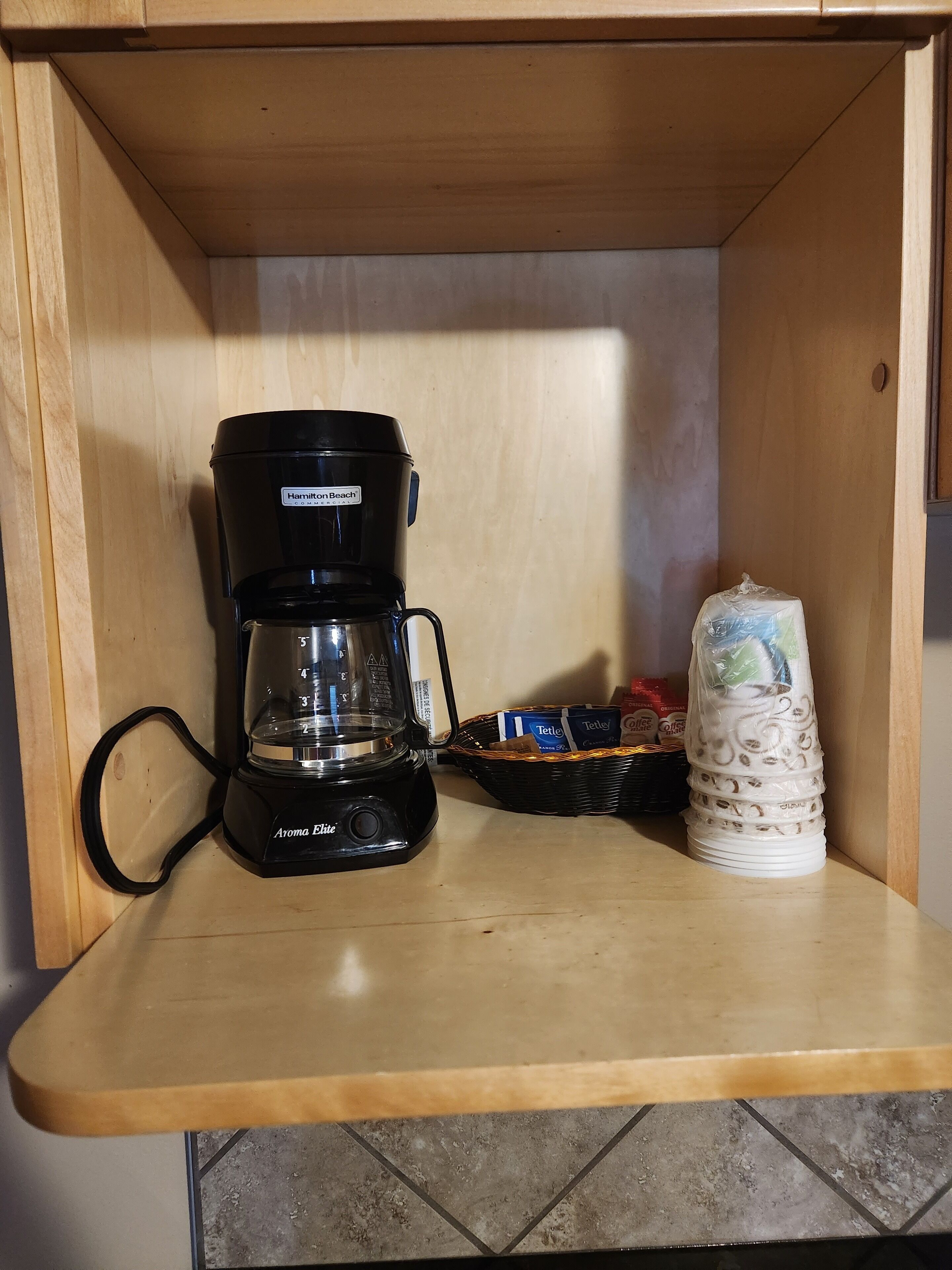 Una piccola mensola di legno con una macchina da caffè nera, un cesto di confezioni Tetra, e una pila di bicchieri bianchi.
