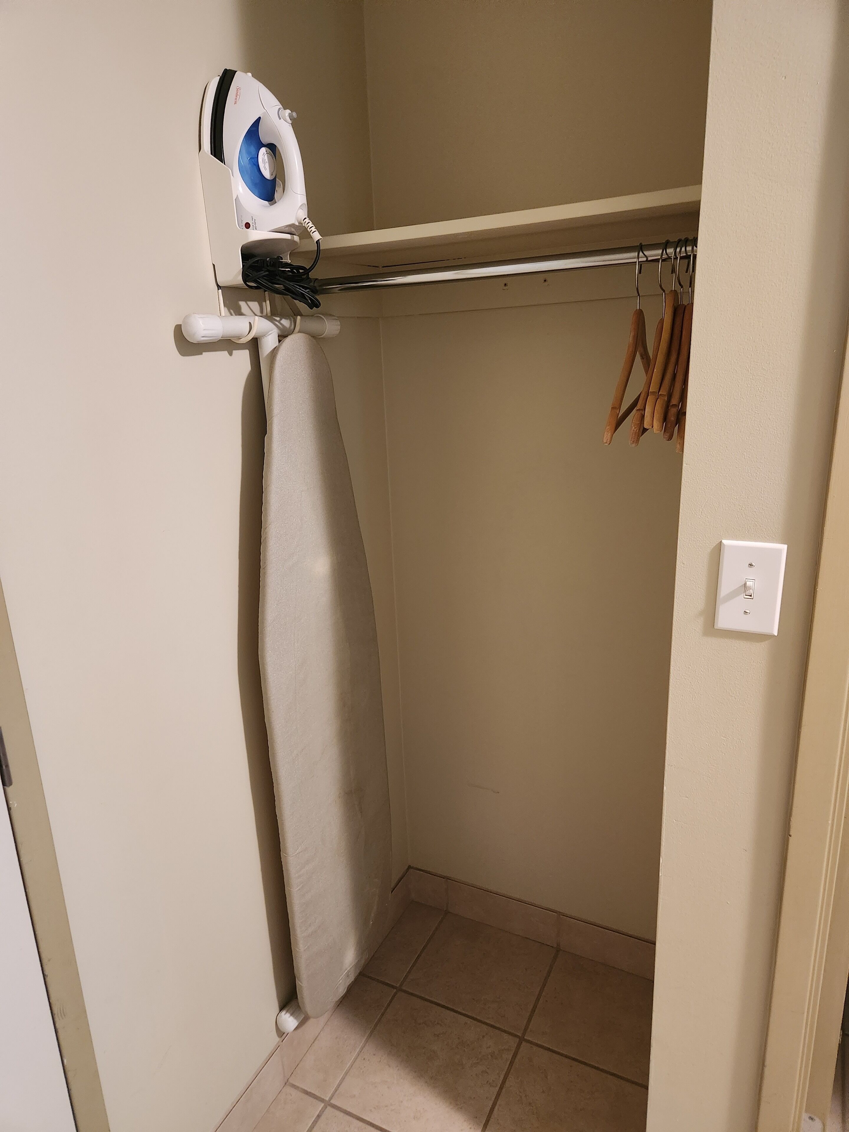 A small, clean room with a wall-mounted ironing board, a clothes rack with wooden hangers, and a light switch on the wall.