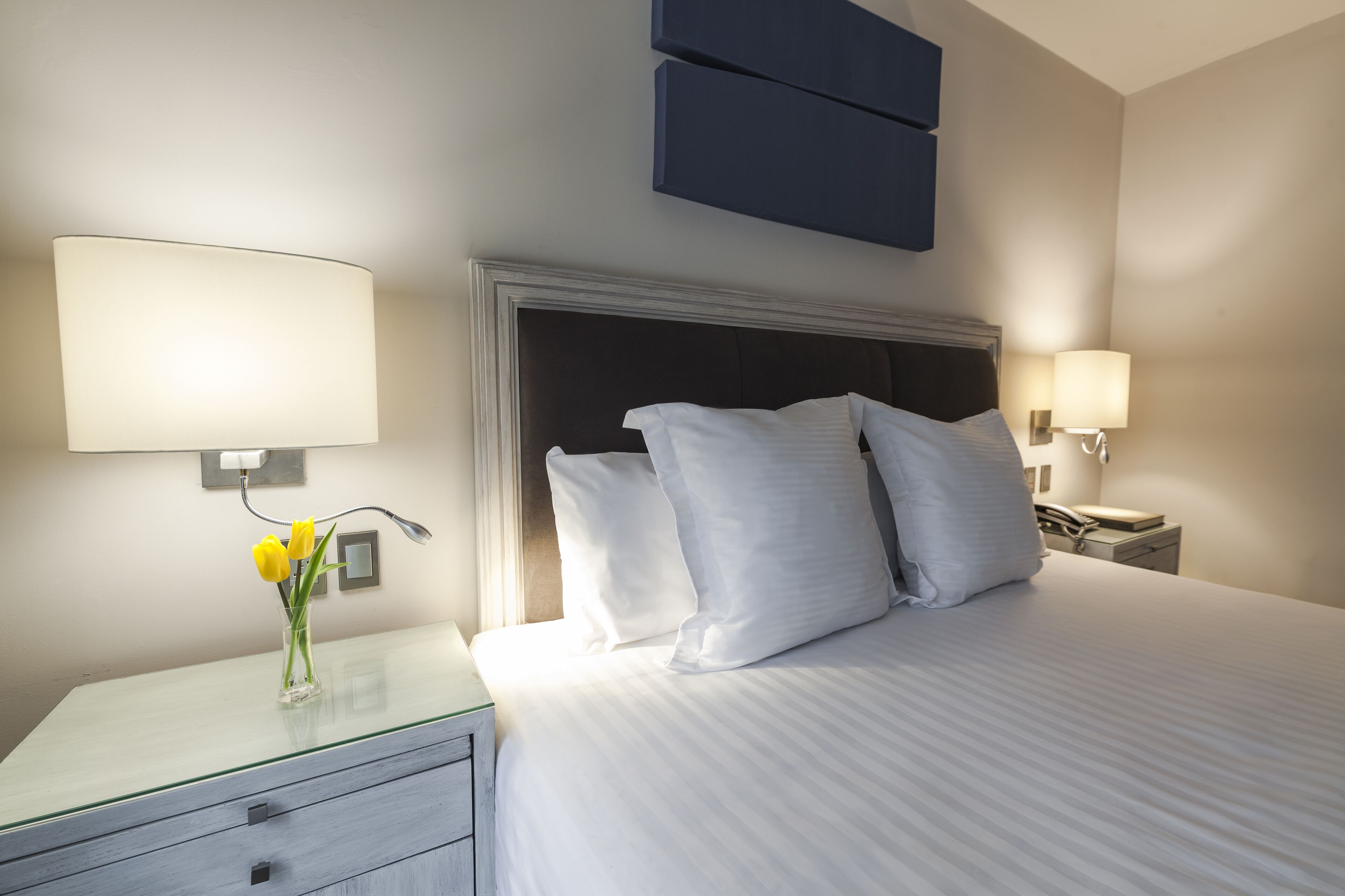 A hotel room with a large bed, two bedside lamps, a nightstand, a glass table with a vase of flowers, and a modern headboard.